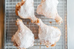 Four chicken drumsticks coated in flour resting on a wire rack before frying.