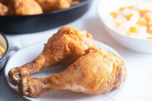 Two golden fried chicken legs on a white plate with sides in the background.