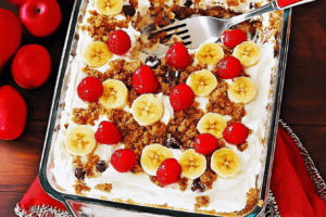 A close-up of a layered no-bake banana split cake topped with bananas, raspberries, and nuts on a white plate.