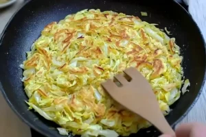 Golden cabbage pancake cooking in a skillet with a wooden spatula.