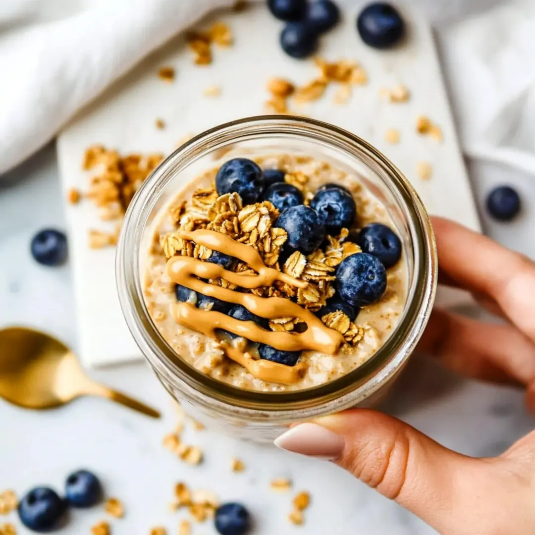 Blended Overnight Oats with Blueberries, Granola, and Peanut Butter Drizzle A hand holding a jar of creamy blended overnight oats topped with blueberries, granola, and a drizzle of peanut butter, with scattered oats and berries in the background.